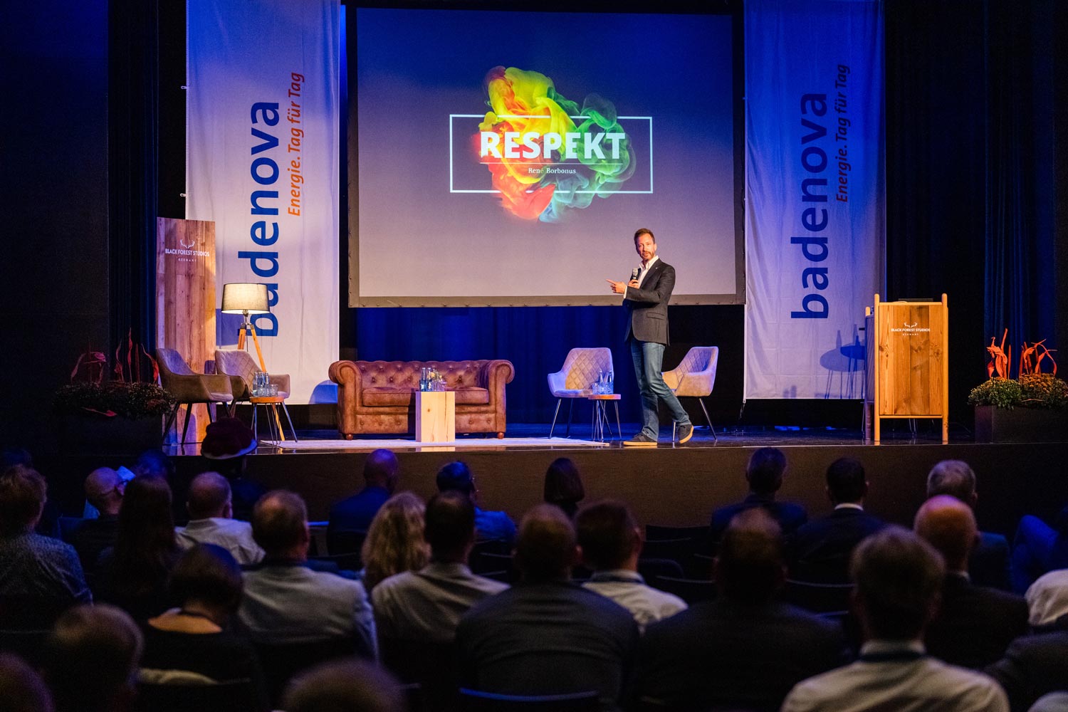 Ein Mann in Anzug und Sneakern steht auf einer großen Bühne und hält eine Keynote-Präsentation. Hinter ihm projiziert eine Leinwand das Wort „RESPEKT“ in bunten Farben. Das Publikum hört aufmerksam zu.