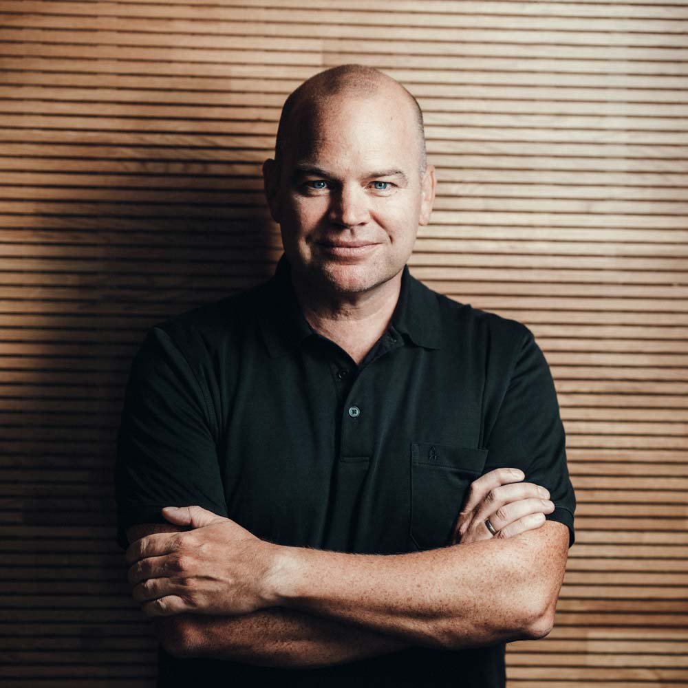 Christian Steiger - Lexware: Mann steht in schwarzem Polo-Shirt mit verschränkten Armen vor einer Holzwand und schaut direkt in die Kamera.