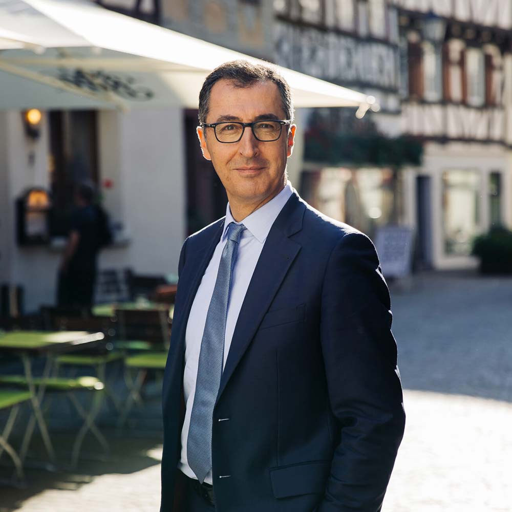Cem Özdemir, Spitzenkandidat der Grünen für die Landtagswahl 2026, ehemaliger Bundesminister: Mann mittleren Alters mit Brille und dunklen Anzug steht auf der Straße vor einem Café mit Blick in die Kamera