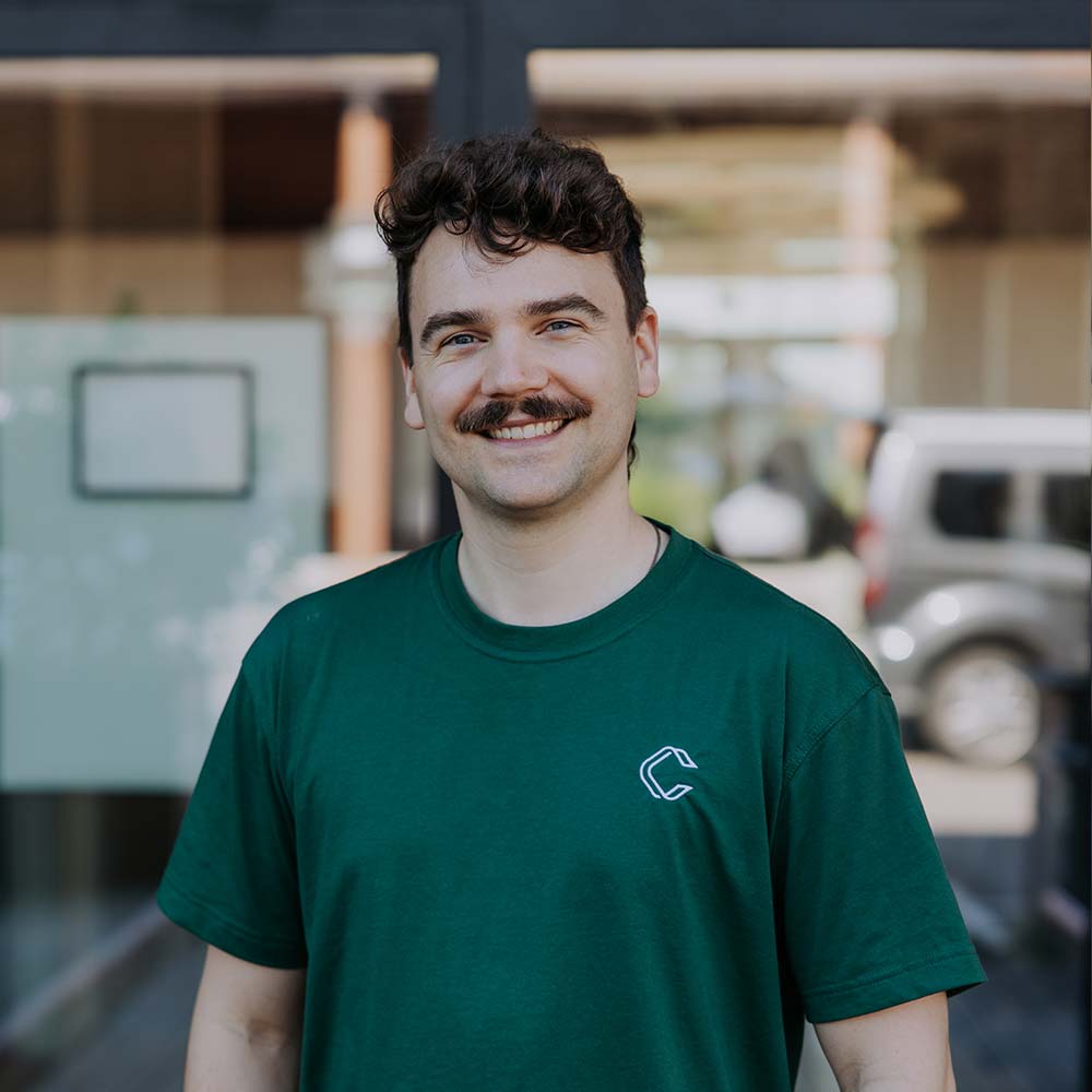 Daniel Kraus, Venture Architect: Junger Mann mit dunkelbraunen Haaren und Schnurrbart in grünem T-Shirt lächelt freundlich in die Kamera