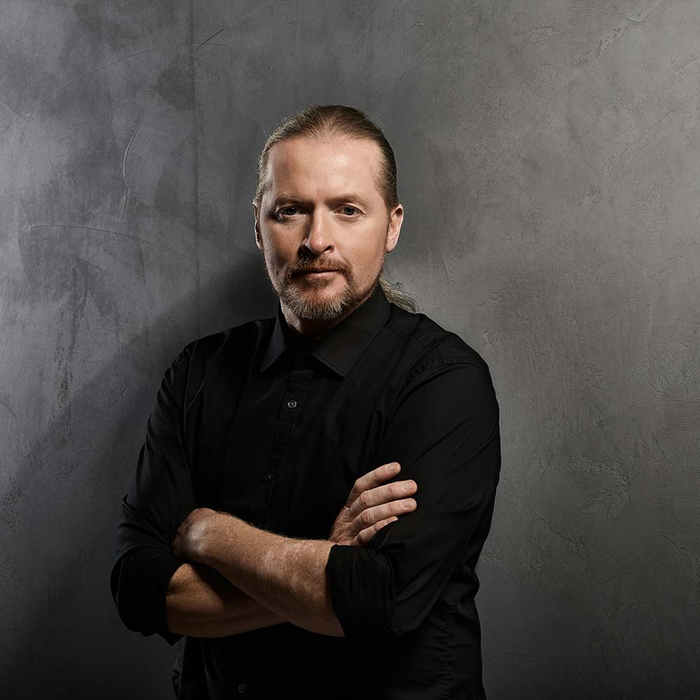 Joey Kelly mit langen, nach hinten gebundenen Haaren und Bart. Er trägt ein schwarzes Hemd und hat die Arme verschränkt vor einer dunklen Wand.