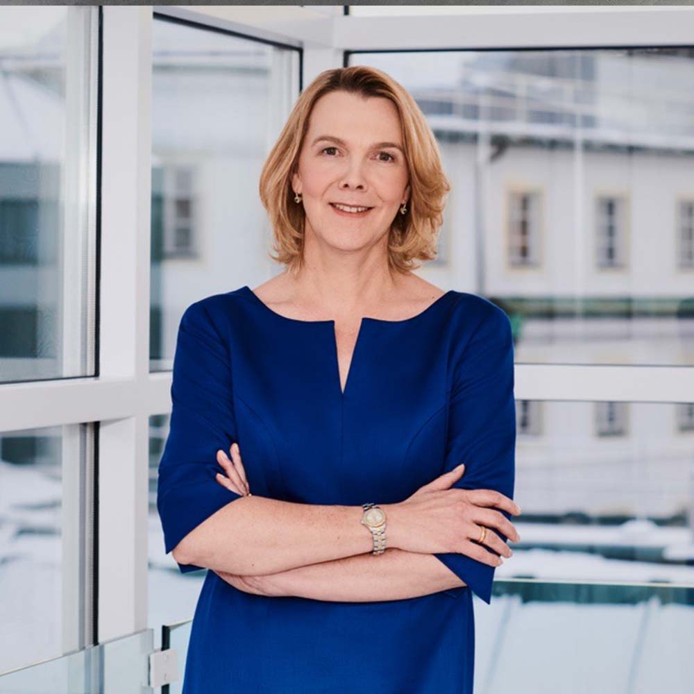 Anne Rethmann hat kurze, dunkelblonde Haare und trägt ein dunkelblaues Langarmshirt. Sie steht vor einer Fensterfront und schaut mit verschränkten Armen und einem Lächeln in die Kamera.