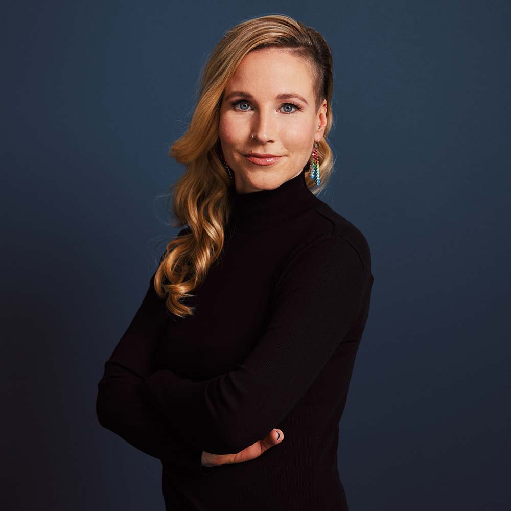 Fränzi Kühne hat blonde, lange Haare und trägt einen schwarzen Rollkragenpullover, bunte Ohrringe und schaut mit leichtem Lächeln in die Kamera.